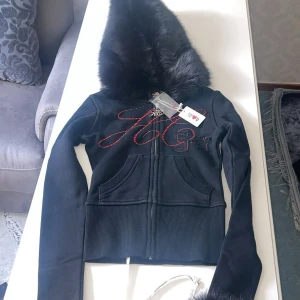 Svart hoodiejacka Hidden Cult pälsdetalj - Svart hoodiejacka från Hidden Cult med stora pälsdetaljer på huva och ärmslut. Jackan har dragkedja framtill, röda strasstenar i logotypen och ribbade muddar. Snygg och edgy stil med croppad passform och långa ärmar.Säljer: Helt ny Hidden Cult hoodie – Storlek XXS   Säljer en helt ny och oanvänd hoodie från populära streetwearmärket Hidden Cult. Modellen är slutsåld på många ställen och perfekt för dig som gillar edgy och unik stil. Svart set med en zip-up hoodie och matchande byxor. Hoodien har