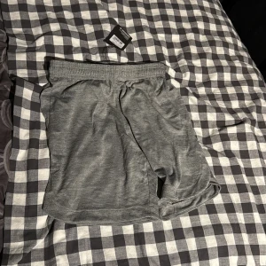 Grå träningsshorts från Energetics - Snygga grå melerade träningsshorts från Energetics, köpta på Intersport. De har elastisk midja med snörning och meshfoder på insidan för extra komfort. Perfekta för gymmet eller löprundan, med en loose passform som ger bra rörelsefrihet.