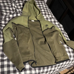 Nike tech Storlek M - Nike hoodiejacka i två nyanser av grönt med svart logga på bröstet. Jackan har hel dragkedja, huva och svarta detaljer längs bröstet och ärmarna. Tillverkad i mjukt material, perfekt för chill eller träning. Snygg och sportig look med praktiska fickor.