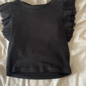 Svart volangtopp från Zara - Supersnygg svart topp från Zara med volangdetaljer på axlarna. Modellen är croppad och har en rund halsringning. Tyget är ribbat och toppen är ärmlös, vilket ger en trendig och cool look. Perfekt att styla med höga jeans eller kjol. På mig som är ungefär 165 cm lång så blir det väldigt mycket magtröja så skulle nog passa bättre på någon mellan 150-160 cm lång