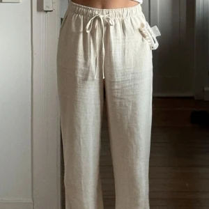 Beiga linnebyxor med snörning från Vila - Säljer ett par beiga linnebyxor från Vila med avslappnad passform och dragsko i midjan. Byxorna har raka ben och är perfekta för en chill och stilren look. Lätta och luftiga, perfekta för varmare dagar.