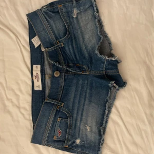 Blå jeansshorts från Hollister - Snygga blå jeansshorts från Hollister med råa kanter och slitna detaljer. Klassisk femficksmodell med knapp och dragkedja framtill samt broderad logga på fickan. Perfekta för varma dagar och en avslappnad stil. Nästana aldrig använda och jättefint skick!