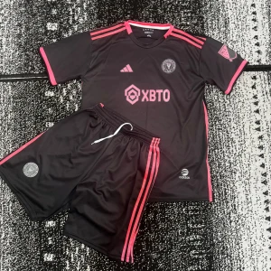 Inter Miami Messi #10 fotbollströja set - Säljer ett Inter Miami fotbollströja och shorts set från Adidas. Tröjan är svart med rosa detaljer, kortärmad och har Messi #10 på ryggen. Klubblogga och sponsortryck på bröstet, klassiska Adidas-ränder på axlar och shorts. Tillverkad i lätt och ventilerande polyester.