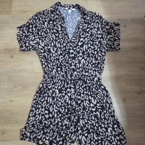 Leopardmönstrad jumpsuit shorts H&M - Snygg leopardmönstrad jumpsuit från H&M i svart och beige. Modellen har korta ben, kort ärm, krage och knappar framtill samt resår i midjan för en skön passform. Perfekt för dig som vill sticka ut med ett djurmönster.
