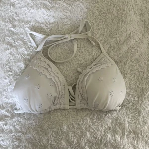 Vit bikiniöverdel med spetsdetaljer - Supersöt vit bikiniöverdel med broderade blommor och spetskant. Triangelformade kupor med knyt i nacken och ryggen. Perfekt för stranden eller poolen, ger en romantisk och fräsch look.