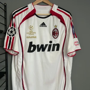 AC Milan Kaka Champions League tröja - AC Milan bortatröja från Champions League-finalen 2007 med Kaka och nummer 22 på ryggen. Vit med röda och svarta detaljer, Adidas-logga, klubbmärke och Bwin-tryck. Kortärmad modell med broderade märken på ärmarna. Perfekt för samlare eller fans. Helt ny med tagg en replika