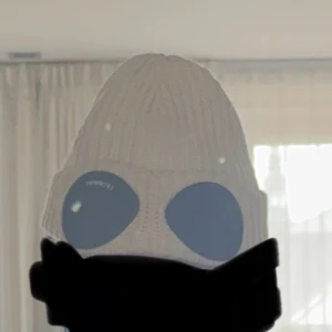 Vit C.P. Company goggle-mössa - Säljer en ikonisk vit ribbstickad mössa från C.P. Company med svarta goggle-linser framtill. Mössan är tillverkad i mjukt stickat material och har en unik streetwear-look som sticker ut. Perfekt för dig som vill ha något extra till vintern.