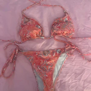 Rosa paisley bikini med knyt - Snygg bikini i rosa med paisleyinspirerat mönster och tunna knytband både upptill och nertill. Triangeltopp och högt skurna trosor ger en trendig look. Perfekt för stranden eller poolen i sommar!