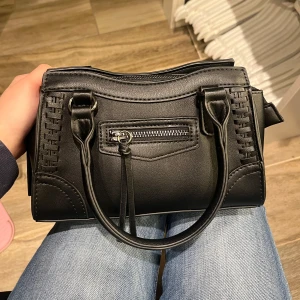 Svart väska - En svart väska från Gina Tricot som aldrig användes, det går att välja om man vill ha axelband eller inte. Finns 2 små fickor med dragkedja och en litet fack i den. Ej använd men tog bort prislappen 
