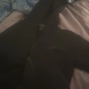 Svart hoodie från Polo Ralph Lauren - Svart hoodie från Polo Ralph Lauren med den klassiska röda broderade loggan på bröstet. Tröjan har huva och är långärmad, perfekt för en chill och stilren look. Tillverkad i mjuk bomull för extra komfort.