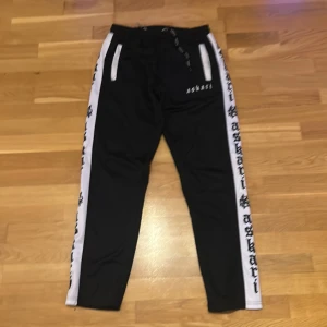 Svarta mjukisbyxor Askari med vita ränder - Svarta mjukisbyxor från Askari med vita sidor och svart text längs benen. Byxorna har snörning i midjan och två fickor framtill med vita detaljer. Perfekta för en chill och sportig stil.