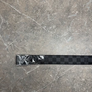 Rutig bälte - Snyggt bälte med ett rutigt mönster i grått och svart. Bältet har en stor L* som spänne och är perfekt för att ge en lyxig touch till din outfit.