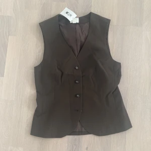 Brun kostymväst med knappar, strl 36 - Snygg brun väst i kostymstil med fyra knappar framtill och v-ringad hals. Figursydd passform som ger en clean och stilren look. Perfekt att styla över en t-shirt eller skjorta för en trendig vibe.