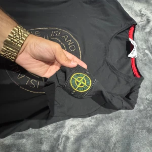 Svart Stone Island t-shirt med emblem 3XL - Svart t-shirt från Stone Island med stort broderat logomärke på bröstet och klassiskt emblem på ärmen. Röd detalj på insidan av kragen. Tillverkad i mjuk bomullsblandning för skön passform. Perfekt för dig som gillar streetwear och vill sticka ut.