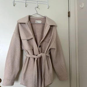 Beige kappa  - Snygg beige overshirt från NLY Trend med bälte i midjan och stora bröstfickor. Materialet har slitits lite. 