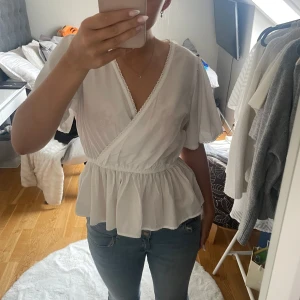 Vit omlottblus från Vero Moda - Supersöt vit omlottblus från Vero Moda med volang nertill och korta, vida ärmar. Blusen har v-ringning med spetskant och resår i midjan. Blusen kan vara lite genomskinlig men är jätte fin ändå. Har endast använt den 1 gång☺️