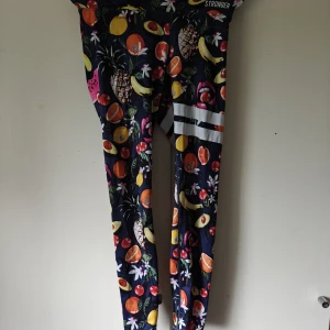 Stronger leggings med fruktmönster - Färgglada leggings från Stronger med tropiskt fruktmönster i gult, rött, grönt och orange på mörkblå botten. Tight passform, vita ränder på ena benet och tillverkade i återvunnen polyester och elastan. Perfekta för träning eller när du vill sticka ut.