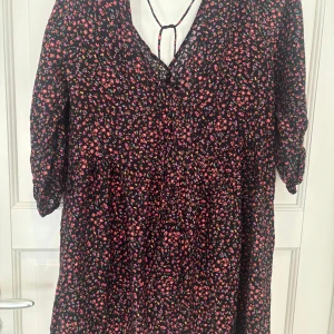 Svart blommig klänning med knyt - Supersöt svart klänning med små blommor i rosa, rött och lila. V-ringad med knyt i ryggen och trekvartsärm. Luftig passform och lätt material, perfekt för vår och sommar. Faller snyggt och har en avslappnad vibe.