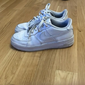 Nike Air Force 1 vita sneakers - Klassiska Nike Air Force 1 sneakers i helvitt skinn med perforerad tå, rund tå och snörning. Snygg och tidlös modell med platt sula och diskret Nike-logga på sidan och hälen. Perfekta för en clean och sportig look.