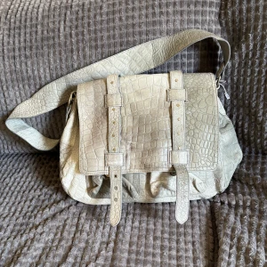 Beige axelväska i krokopräglat skinn - Säljer en grå  axelväska med krokodilpräglat mönster och bred axelrem. Väskan har två spännen framtill och rymligt huvudfack. Perfekt för dig som vill ha en unik och stilren väska med cool struktur.