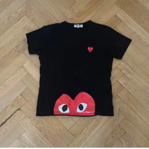 Svart Comme des Garçons Play t-shirt - Svart t-shirt från Comme des Garçons Play med ikoniskt rött hjärta med ögon på bröstet och ett stort hjärta med ögon tryckt längst ner. Klassisk passform i storlek L.