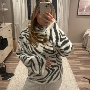 Zebramönstrad fluffig stickad tröja - Supermysig stickad tröja med zebramönster i svart och vitt. Tröjan har hög hals, lång ärm och är riktigt fluffig och skön. Perfekt för dig som vill sticka ut med en cool och trendig look. Passar till både jeans och mjukisbyxor.