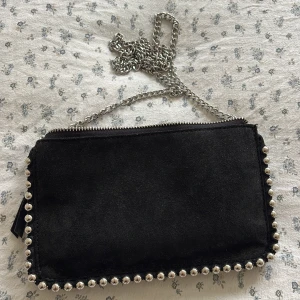 Svart axelväska med kedja och nitar - Snygg svart axelväska i mocka med silverfärgad kedja och coola nitar längs kanterna. Väskan har dragkedja upptill och flera fack inutiz Tryck på köp nu!