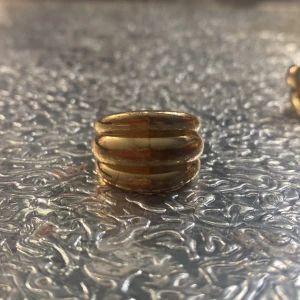Chunky guldig ring med räfflad design - Säljer en chunky ring i guldton med cool räfflad och rundad form. Ringen har tre upphöjda band som ger en statement-look och passar perfekt för dig som gillar att sticka ut. Ingen märkning syns på insidan. Perfekt accessoar för att lyfta din outfit.