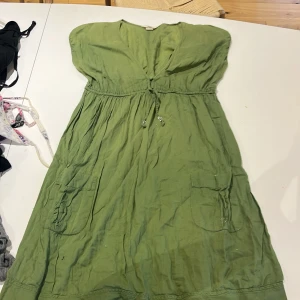 Grön bomullsklänning från L.O.G.G. - Snygg grön klänning från L.O.G.G. med v-ringning, knyt i midjan och två stora fickor framtill. Tillverkad i mjuk bomull, perfekt för varma dagar. Avslappnad passform och kort ärm för en chill vibe.