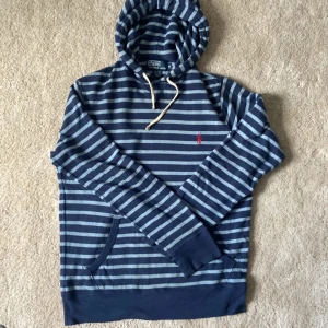 Blå- och grårandig hoodie från Polo Ralph Lauren - Säljer en blå- och grårandig hoodie från Polo Ralph Lauren med huva och dragsko. Tröjan har en broderad röd logga på bröstet och en praktisk ficka framtill. Perfekt för en avslappnad stil.