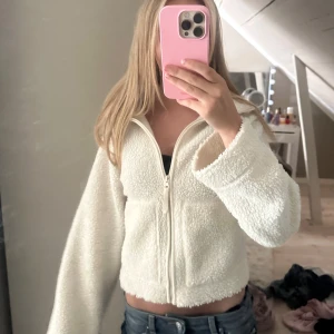 Vit teddyjacka med dragkedja - Supermysig vit teddyjacka med croppad passform och lång ärm. Jackan har en hel dragkedja framtill och är gjord i fluffigt fleece-material som känns riktigt skönt mot huden. Perfekt för dig som gillar en chill och trendig stil.