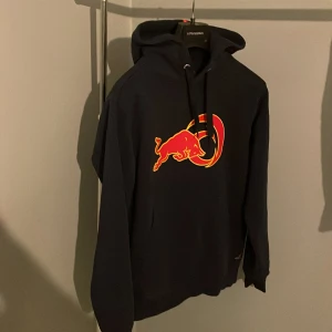 Svart Red Bull hoodie från Sail Racing - Svart hoodie från Sail Racing x Alinghi Red Bull Racing i storlek medium. Stor röd och gul Red Bull-logga på bröstet, huva med snörning och klassisk känguruficka. Skön passform och perfekt för dig som gillar sportiga och snygga streetwear-plagg.