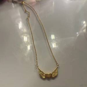 Guldhalsband med hjärta och vingar - Snyggt halsband i guld med ett hängsmycke som föreställer ett hjärta med vingar på sidorna. Kedjan är tunn och elegant, perfekt för dig som gillar detaljer med lite edge och symbolik. Passar till alla tillfällen och ger din outfit en unik touch.
