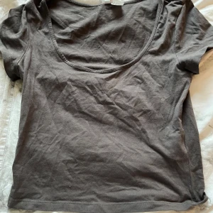 Mörkgrå croppad topp från H&M Divided - En enkel och stilren mörkgrå croppad topp från H&M Divided. Modellen har korta ärmar och en djup rundad halsringning. Tillverkad i mjuk bomull som är skön mot huden. Perfekt att matcha med jeans eller kjol för en chill look.