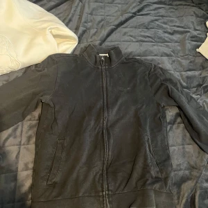 Svart och vit overshirt från EA7 - Två stilrena overshirts från EA7, en i svart och en i vit. Båda har dragkedja framtill, ståkrage och fickor på sidorna. EA7-loggan syns diskret på bröstet. Materialet är mjukt och känns som bomull, perfekt för en chill och sportig look. 2 för 400 kr