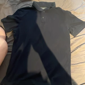 Mörkblå pikétröja från Dressmann S - Klassisk svart pikétröja från Dressmann i storlek S. Tröjan har korta ärmar, krage och två knappar framtill. Materialet är mjuk bomull som känns skönt mot huden. Perfekt för dig som gillar enkel och stilren look.