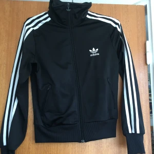 Svart Adidas träningsjacka S - Svart Adidas vindjacka med klassiska vita ränder längs ärmarna och stort vitt trefoil-tryck på ryggen. Jackan har hög krage, dragkedja framtill och två fickor med dragkedja. Materialet är polyester och passformen är normal. Perfekt för en sportig streetstyle-look.