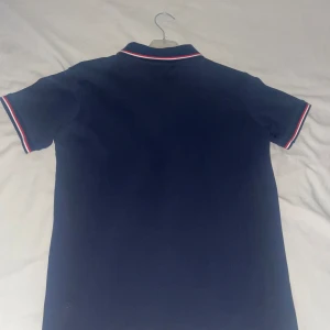 Mörkblå Moncler pikétröja - Snygg mörkblå pikétröja från Moncler med klassisk krage och korta ärmar. Tröjan har vita och röda ränder på krage och ärmslut samt Moncler-logga på bröstet. Helt ny