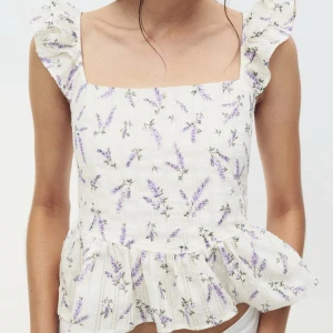 Vit blommig topp med volang detaljer  - Supersöt blus i vit bomull med lavendelfärgat blommönster och volangärmar. Toppen har fyrkantig ringning, knytning i ryggen och lätt rynkad nederkant. Tyvärr förliten för mig, så den är fortfarande i nyskick!