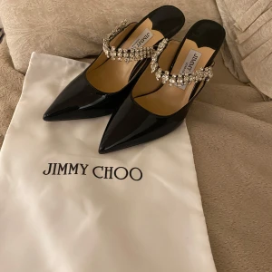 Svarta pumps med strass från Jimmy Choo - Säljer ett par svarta pumps från Jimmy Choo med spetsig tå och hög klack. Skorna har en glansig finish i skinn och är dekorerade med en lyxig strassrem över foten. Perfekta för dig som vill sticka ut med elegans och bling. Passar storlek 37, 37,5 och 38. Oanvända kommer endast med dustbagen
