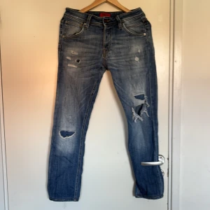 Blå ripped jeans från Jack & Jones - Snygga blå jeans från Jack & Jones med slitna detaljer och stora hål på benen för en cool, edgy vibe. Klassisk femficksmodell med normal passform och midwaist. Jeansen har en tvättad look och är gjorda i mjukt denimtyg med dragkedja och knapp.
