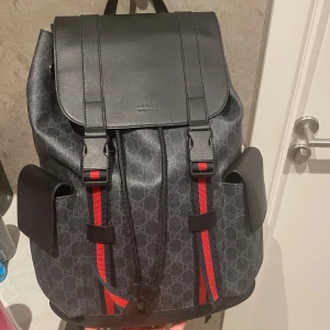 Gucci ryggsäck med GG-mönster - Snygg ryggsäck från Gucci i mörkgrått med klassiskt GG-mönster och svarta detaljer i skinn. Väskan har två sidofickor, lock med spännen och röda/blå ränder framtill. Perfekt för dig som vill ha en lyxig och praktisk väska. Fler bilder kan fås och den har inga defekter då jag har tagit hand om det väldigt väl och den har endast hängt på en vägg som prydnad. Hör gärna av dig! Vid lila minsta fundering. Väskan är själv klart äkta! Pris kan diskuteras vid snabb affär👍