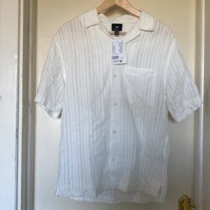 Vit randig skjorta från H&M - Snygg vit skjorta från H&M med diskreta vertikala ränder. Regular fit med korta ärmar, klassisk krage och bröstficka. Perfekt för varma dagar och chill stil. Tillverkad i lätt bomullsmaterial som andas. Knappar framtill för enkel på- och avtagning.