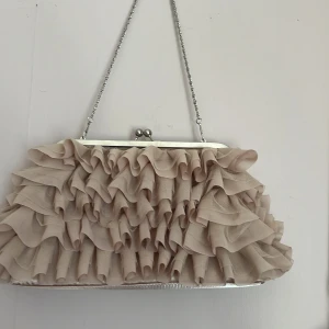 Beige kuvertväska med volanger från Tiamo - Säljer en beige kuvertväska från Tiamo med snygga volanger i tyg och silverfärgad metallram. Väskan har en kedja för att hänga över axeln och ett innerfack med dragkedja. Perfekt för att lyfta din outfit med en unik detalj.