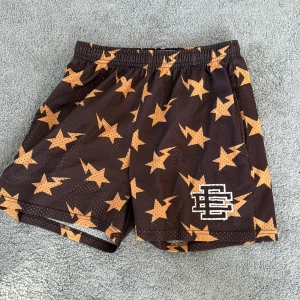  EE erica Emanuel shorts  - Snygga bruna meshshorts från EE med orangea stjärnor och blixtar över hela tyget. Elastisk midja med vit snörning och broderad EE-logga på benet. Perfekta för dig som gillar streetwear och vill sticka ut med coolt mönster.