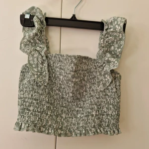 Grön blommig topp  - Grän topp med vita blommor. Ursprungligen från Shein. Magtröja så den är kort. Det står att den är för 14 åringar men den är liten i storleken så jag skulle nog säga 11-12 år så kanske storlek 152 men det är olika från person till person. 