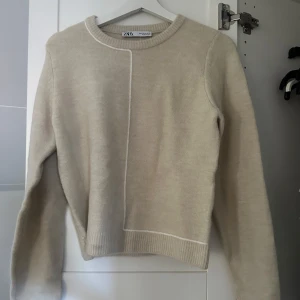 Beige stickad tröja från Zara - Snygg beige stickad tröja från Zara med rund hals och långärmad design. Tröjan har en cool vit söm som går vertikalt framtill och ribbade muddar vid ärmslut och nederkant. Perfekt för dig som gillar stilrena och mjuka plagg.