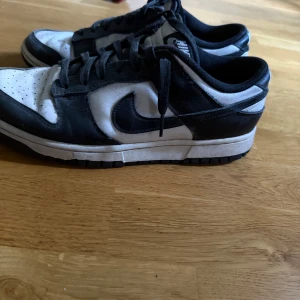 Nike Dunk svartvita sneakers - Säljer ett par Nike Dunk sneakers i svart och vitt med klassisk låg siluett. Skorna har snörning, rund tå och perforerad tåbox för extra ventilation. Swoosh-loggan syns tydligt på sidan och ovandelen är i skinn. Perfekt för dig som gillar streetwear och vill ha en ikonisk look.