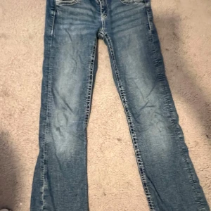 Blå jeans från Gina  - Säljer ett par blå jeans med snygga broderade detaljer på bakfickorna och silverfärgade knappar. Jeansen är slitna där vid bak fickan men den syns inte igenom (har sytt igenom hållet)