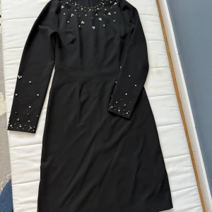 Svart klänning med pärldetaljer - Snygg svart långklänning med rund hals och långa ärmar, dekorerad med pärlor vid halsringningen och på ärmarna. Klänningen har en slits baktill för extra rörelsefrihet och är tillverkad i ett mjukt, slätt material som ger en elegant siluett.